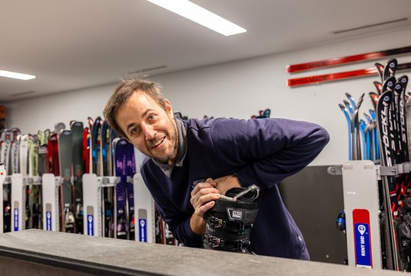 Ein Mitarbeiter in einem Skigeschäft passt einem Kunden einen Skischuh an; im Hintergrund sind Regale mit verschiedenen Skischuh-Modellen zu sehen.