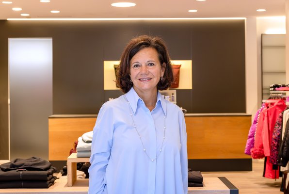 Eine lächelnde Frau steht in der Modeabteilung eines Sportgeschäfts und blickt direkt in die Kamera.