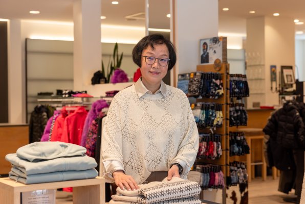 Lächelnde Mitarbeiterin legt in der Modeabteilung einen Pullover zusammen und blickt in die Kamera.