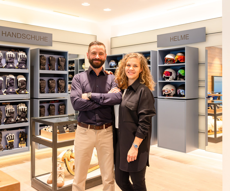 Die Geschäftsführer Michi und Riccarda Habegger stehen gemeinsam in der Modeabteilung ihres Skigeschäfts; im Hintergrund sind blaue Regale mit Skihelmen und Handschuhen zu sehen.