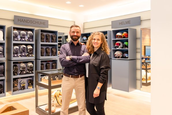 Die Geschäftsführer Michi und Riccarda Habegger stehen gemeinsam in der Modeabteilung ihres Skigeschäfts; im Hintergrund sind blaue Regale mit Skihelmen und Handschuhen zu sehen.