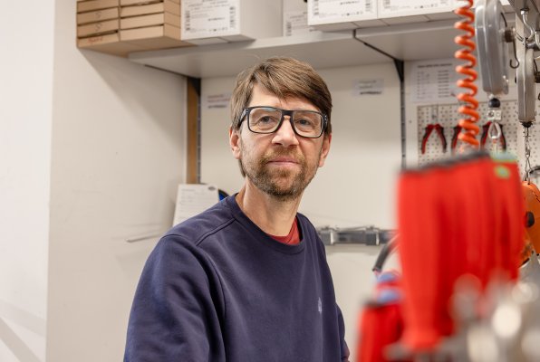 Ein Mitarbeiter blickt lächelnd in die Kamera; im Hintergrund ist eine Werkstatt mit Werkzeugen und Ausrüstung zu sehen.