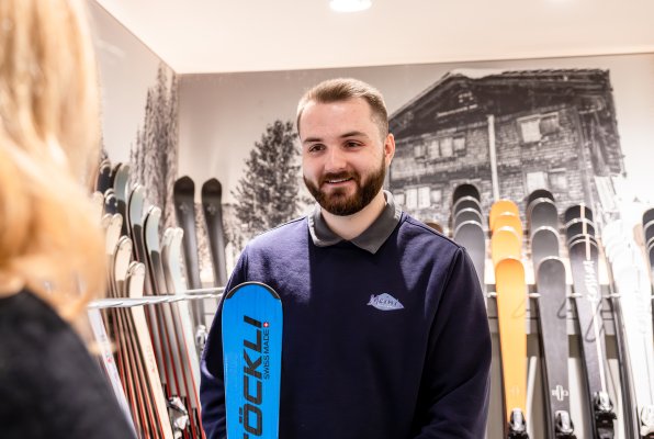 Ein lächelnder Mitarbeiter in einem Sportgeschäft berät eine Kundin und hält dabei einen blauen Ski der Marke Stöckli in der Hand.
